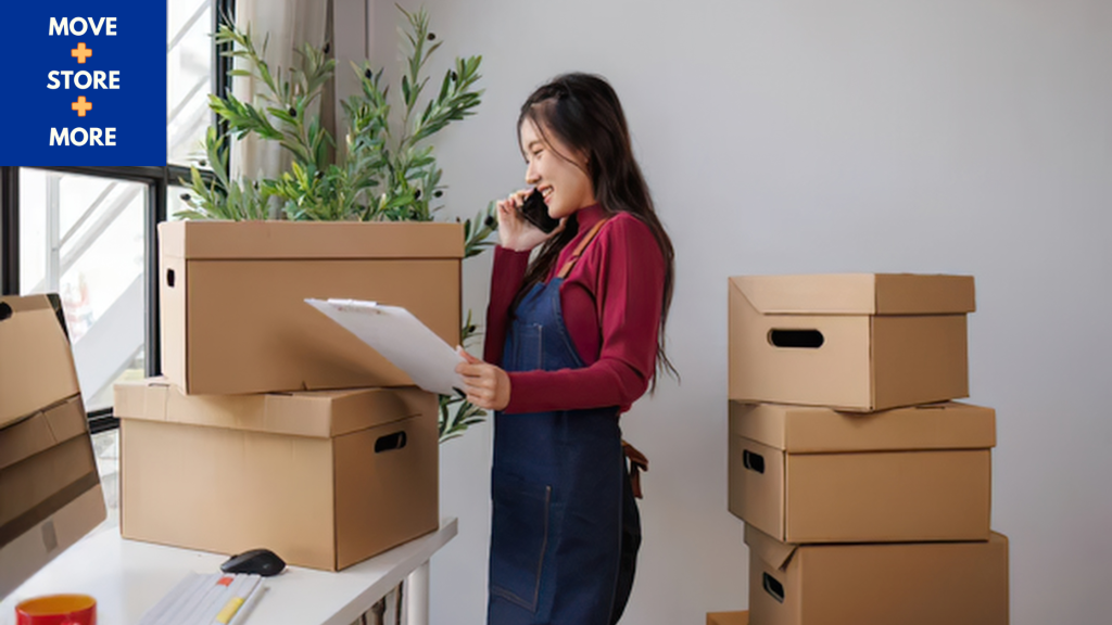 Image of a woman holding a board beside packing boxes - Move Store & More How to Save Money Before Moving House Blog Image